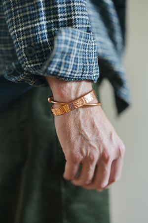 Kopero Kobberarmbånd Rund, om håndleddet over en ternet skjorteærme, sammen med et hamret armbånd